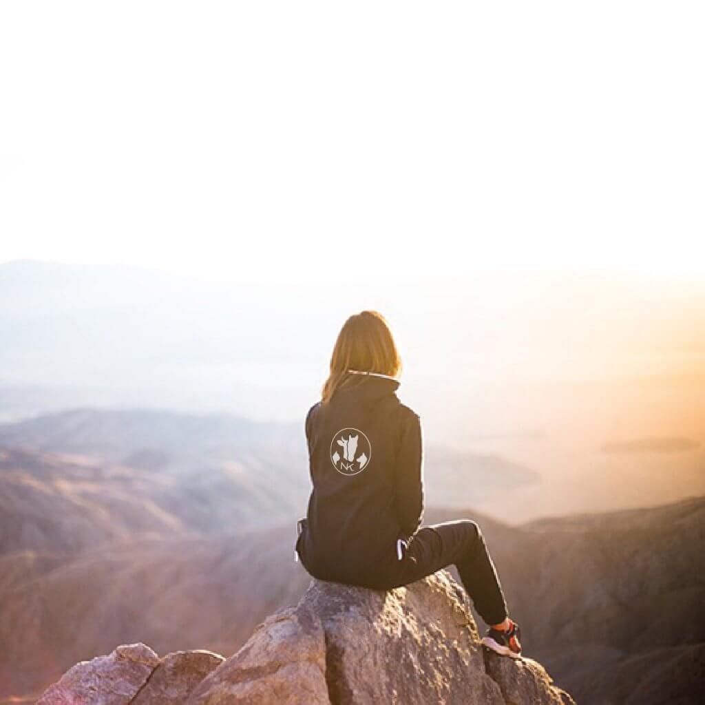 Femme assise sur un rocher surplombant un paysage montagneux, portant une veste avec le logo de Natacha Kozak au dos.